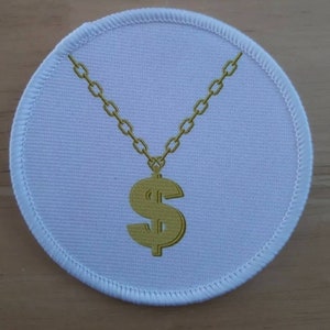 Può includere: Un cerotto rotondo bianco con una catena dorata e un ciondolo a forma di dollaro. La catena è avvolta attorno alla parte superiore del cerotto e il ciondolo pende al centro.