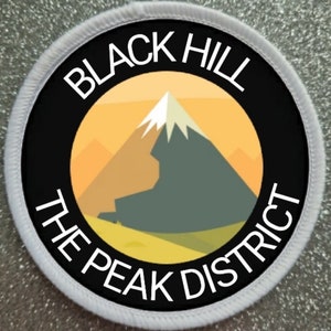Op de afbeelding: Een witte ronde patch met een zwarte cirkel in het midden. De zwarte cirkel heeft witte tekst die "BLACK HILL THE PEAK DISTRICT" luidt en een afbeelding van een berg met een besneeuwde top in het midden.