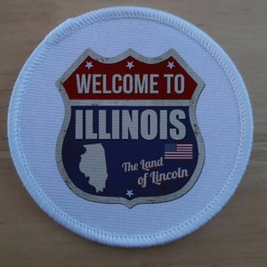 Op de afbeelding: Een witte ronde patch met een blauwe, rode en witte afbeelding van een vintage-snelweg bord met de tekst "Welcome to Illinois, The Land of Lincoln". Het bord heeft een witte omtrek en sterren bovenaan.