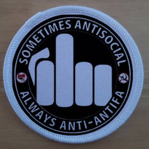 Può includere: Una toppa rotonda con bordo bianco e la scritta "SOMETIMES ANTISOCIAL" e "ALWAYS ANTI-ANTIFA" in un cerchio nero. Il centro presenta un gesto della mano bianca con il dito medio alzato. Piccoli simboli rossi e bianchi su entrambi i lati.
