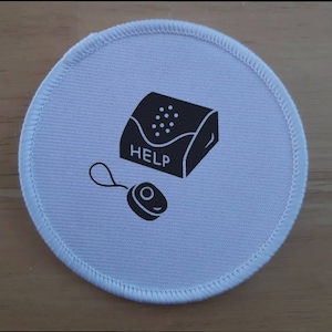 Peut inclure: Patch rond en tissu blanc avec une illustration en noir et blanc d'un système d'alarme personnel. Le système d'alarme a un bouton qui dit "HELP" et un petit cordon noir.
