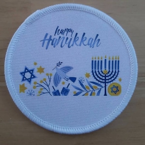 May include: A round white patch with a stitched border. The patch features the words "happy Hanukkah" in blue script. Below the text are illustrations of a dove, dreidels, a menorah, and the Star of David in blue and yellow.