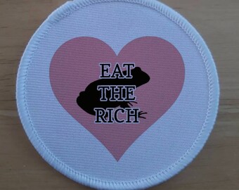 May include: A white round patch with a pink heart in the center. The heart has a black silhouette of a frog and the text "EAT THE RICH" in black.