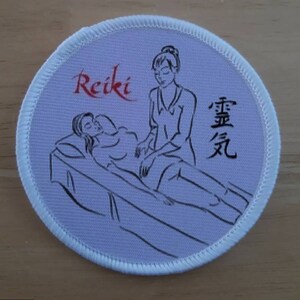May include: White round patch with a black and white illustration of a Reiki practitioner giving a treatment to a person lying on a table. The text "Reiki" is written in red cursive and the text "霊気" is written in black cursive.