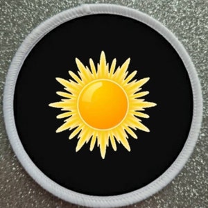 Könnte beinhalten: Runder Aufnäher mit einem leuchtend gelben Sonnendesign auf schwarzem Hintergrund. Die Sonne hat eine gelbe Mitte und spitze Strahlen. Der Aufnäher ist mit einem weißen, genähten Rand versehen.