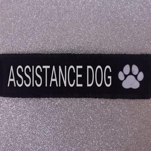May include: Black fabric patch with white text that reads "ASSISTANCE DOG" and a white paw print.
