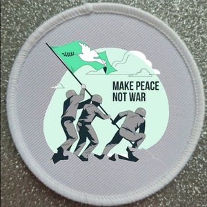 Può includere: Un cerotto rotondo bianco con una bandiera verde che presenta una colomba bianca e il testo "MAKE PEACE NOT WAR". Tre figure in grigio stanno tirando l'asta della bandiera.
