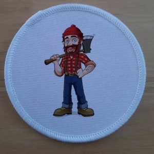 Puede incluir: Un parche redondo blanco con una imagen de dibujos animados de un leñador que lleva una camisa de cuadros rojos y negros, un sombrero rojo y pantalones vaqueros azules. Lleva un hacha sobre el hombro y tiene barba.