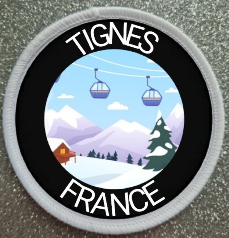 Peut inclure: Un patch circulaire blanc et noir avec le texte "Tignes France" et une image de dessin anim&eacute; d'une sc&egrave;ne de montagne enneig&eacute;e avec des remont&eacute;es m&eacute;caniques et un petit chalet.