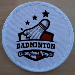 May include: White round patch with a badminton shuttlecock design. The shuttlecock is white and gray with red accents. The text "Badminton Champions League" is printed in black on the patch.