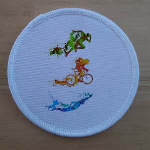 May include: A white round patch with a colorful design of three athletes. The top athlete is running, the middle athlete is cycling, and the bottom athlete is swimming. The design is in a watercolor style.