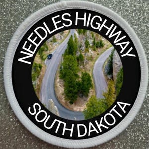 Könnte beinhalten: Runder Aufnäher mit einer malerischen Ansicht des Needles Highway in South Dakota. Das Bild zeigt eine kurvenreiche Straße durch eine bergige Landschaft mit grünen Bäumen. Der Text "NEEDLES HIGHWAY SOUTH DAKOTA" ist in weißen Buchstaben auf schwarzem Hintergrund gedruckt.