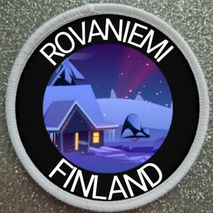 Puede incluir: Un parche redondo blanco con texto negro que dice "ROVANIE MI FINLAND". El centro del parche presenta una imagen de dibujos animados de un paisaje nevado con una casa y una aurora boreal púrpura en el cielo.