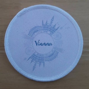 May include: White round fabric patch with a blue outline and a blue silhouette of the Vienna skyline. The text "Vienna" is printed in blue in the center of the patch.