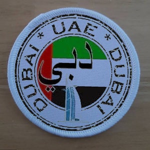 Könnte beinhalten: Ein weißer runder Patch mit schwarzem Rand und dem Text "DUBAI * UAE * DUBAI" am Rand. Die Mitte des Patches zeigt die Flagge der Vereinigten Arabischen Emirate mit dem arabischen Wort für "Dubai" in Schwarz.