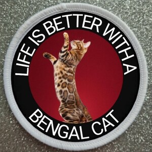 May include: A white round patch with a black border and red center. The patch has the text "Life is better with a Bengal Cat" in white lettering around the edge. A Bengal cat is pictured in the center of the patch.