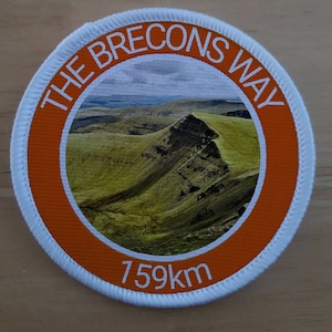 Op de afbeelding: Een ronde witte en oranje patch met de tekst "The Brecons Way" in witte letters rond een foto van een groene bergketen. De tekst "159km" is in wit gedrukt aan de onderkant van de patch.
