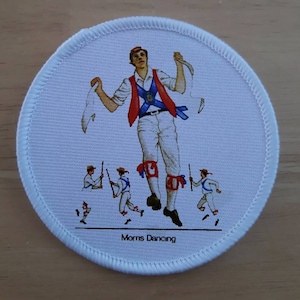 May include: A white round patch featuring a colourful illustration of a Morris dancer in a white shirt, red waistcoat, and white trousers. The dancer is holding a stick and a white handkerchief. The patch also includes four smaller Morris dancers in the bottom half of the image. The text "Morris Dancing" is printed at the bottom of the patch.