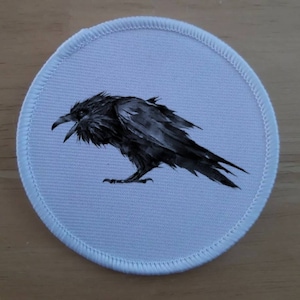 May include: A white round patch with a black and white illustration of a raven. The raven is facing left and has its beak open.