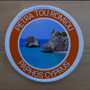May include: A white round patch with an orange border. The patch features a photo of the Petra tou Romiou sea stacks in Paphos, Cyprus. The text "Petra tou Romiou" and "Paphos Cyprus" are printed in white around the photo.