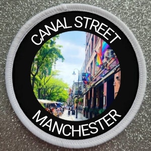 May include: A white round patch with black text that reads "CANAL STREET MANCHESTER". The patch has a circular image of a street scene with buildings and trees in the centre.
