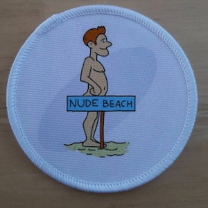 May include: A round, white patch with a cartoon illustration of a nude person standing in front of a sign that reads "NUDE BEACH". The person has red hair and is standing on a patch of green grass.