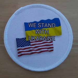 May include: A white round patch with the text "WE STAND WITH UKRAINE" printed in black. The patch features the American flag and the Ukrainian flag.