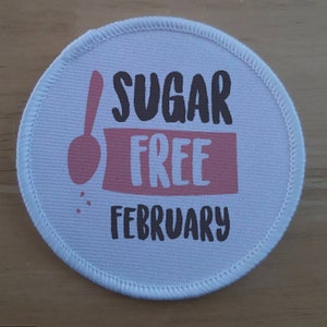 May include: A round, white patch with a stitched border. The patch features the words "SUGAR FREE FEBRUARY" in black and pink lettering, with a pink spoon graphic.