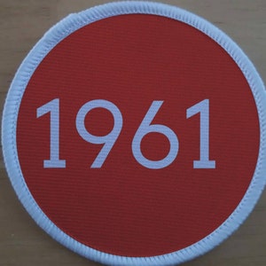 Könnte beinhalten: Ein roter runder Patch mit weißem Rand und der Zahl 1961 in weiß gedruckt.
