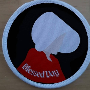 May include: A white and red embroidered patch featuring a silhouette of a woman wearing a white bonnet and red dress. The text "Blessed Day" is embroidered in white on the red dress.