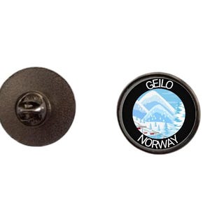 May include: A round metal pin featuring a black border with the white text GEILO NORWAY. The center displays a light blue illustration of snow-covered mountains, pine trees, and a ski lift with red chairs against a pale blue sky.