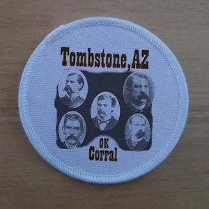 Op de afbeelding: Een witte ronde patch met bruine tekst die "Tombstone, AZ" en "OK Corral" luidt. De patch toont vijf zwart-witportretten van mannen.