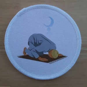 Puede incluir: Parche redondo de tela blanca con una luna creciente azul y una representación de una persona arrodillada en oración sobre una alfombra de oración marrón.