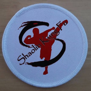 Peut inclure: Patch rond blanc avec un logo Shaolin Kung Fu noir et rouge. Le logo représente un 'S' stylisé avec une silhouette rouge d'une personne donnant un coup de pied en position de combat.
