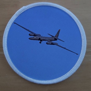 Pode incluir: Um patch redondo com um avião preto em voo contra um fundo azul claro. O patch tem uma borda branca com uma borda costurada. O avião tem asas longas e uma forma distinta.