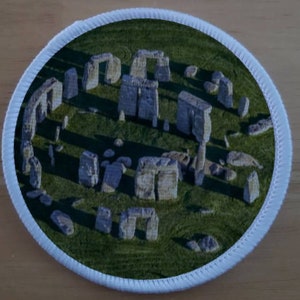 Pode incluir: Remendo redondo com uma imagem detalhada de Stonehenge, um monumento pré-histórico. A imagem mostra o círculo de pedra em um campo gramado verde, cercado por uma borda branca costurada. Para costurar em roupas ou bolsas.