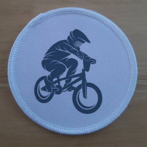 Peut inclure: Un écusson rond blanc avec une silhouette noire d'une personne à vélo BMX. La personne porte un casque et des gants.