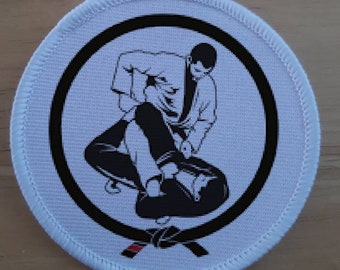 May include: A white circular patch with a black border and a black and white illustration of two people in a martial arts grappling position. A black belt with a red stripe is shown at the bottom of the patch.