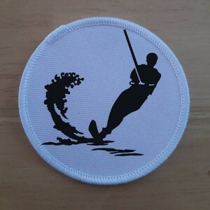 May include: White round patch with a black silhouette of a person water skiing. The person is holding a ski rope and is being pulled by a boat. The water is shown as a black wave behind the skier.