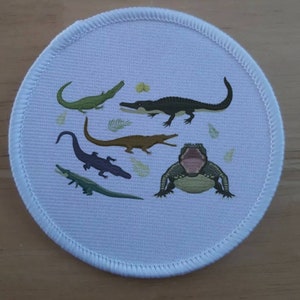May include: A white round patch featuring a colourful design of five alligators. The alligators are in various poses, including one with its mouth open. The patch has a white border with a stitched edge.