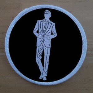 Op de afbeelding: Een witte omtrek van een persoon in een pak tegen een zwarte achtergrond. De persoon staat met zijn handen voor zich gevouwen. De afbeelding is rond en heeft een witte rand.