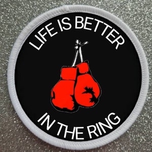 May include: A white circular patch with a black circle in the center. The black circle has the text "Life is better in the ring" in white lettering. There are two red boxing gloves hanging from the top of the circle.