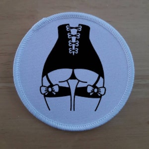 May include: A white round patch with a black silhouette of a person wearing a corset and garters with bows. The person is facing away from the viewer.