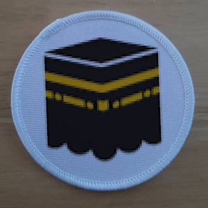 May include: A round, white patch with a stitched border. The patch features a black cube with gold accents, likely representing the Kaaba. The design is centered on the patch, creating a simple yet symbolic image.