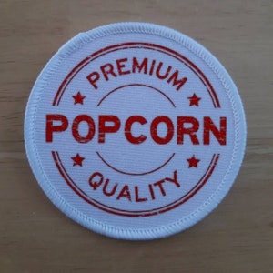 Peut inclure: Écusson rond en tissu blanc avec des coutures rouges autour du bord. L'écusson a un cercle rouge avec le texte "PREMIUM POPCORN QUALITY" dans une police d'écriture usée.