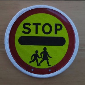 May include: A round, white-bordered sign with a red border and a yellow centre. The sign says "STOP" in black letters and has a black horizontal bar across the centre. There are two stick figures of children running in the lower half of the sign.