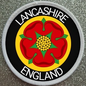 Puede incluir: Un parche circular blanco con un círculo negro en el centro. El círculo negro tiene un círculo amarillo con una rosa roja en el centro. La rosa tiene un centro verde y pétalos amarillos. El texto "LANCASHIRE" está escrito en blanco alrededor de la parte superior del círculo negro y el texto "ENGLAND" está escrito en blanco alrededor de la parte inferior del círculo negro.