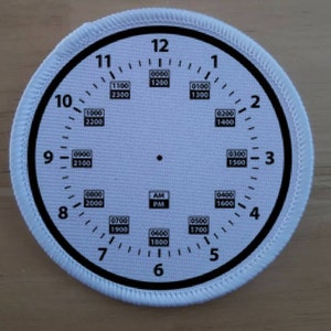 May include: White circular patch with a black clock face design. The clock face features black numerals, minute markers, and military time notations. The patch has a white stitched border and includes "AM" and "PM" indicators.