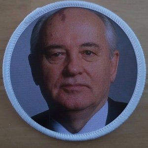 May include: A white circular patch with a white border and a photo of a man with gray hair and a white shirt.