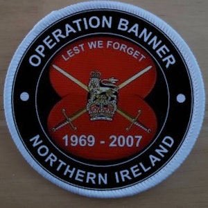 May include: A black and white circular patch with the text "Operation Banner" and "Northern Ireland" around the edge. The center of the patch features a red poppy with crossed swords and a crown. The text "Lest We Forget" and "1969 - 2007" are also included.